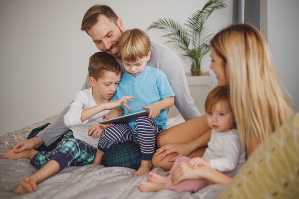A family on a bed, two children leaning over a tablet with their parents.