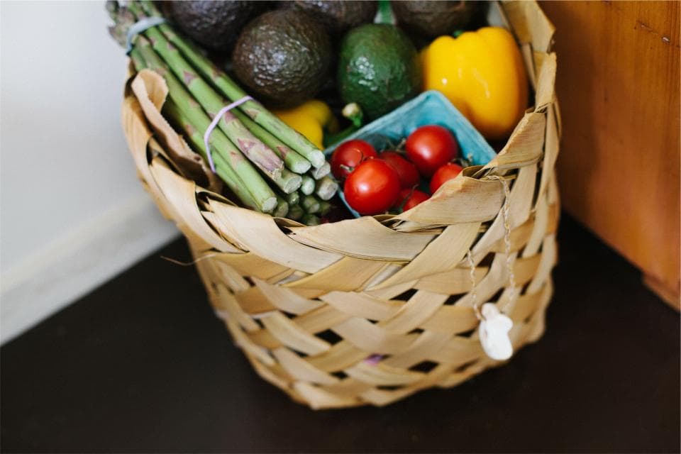 A woven basket full of asparagus, avocados, tomatoes and peppers.