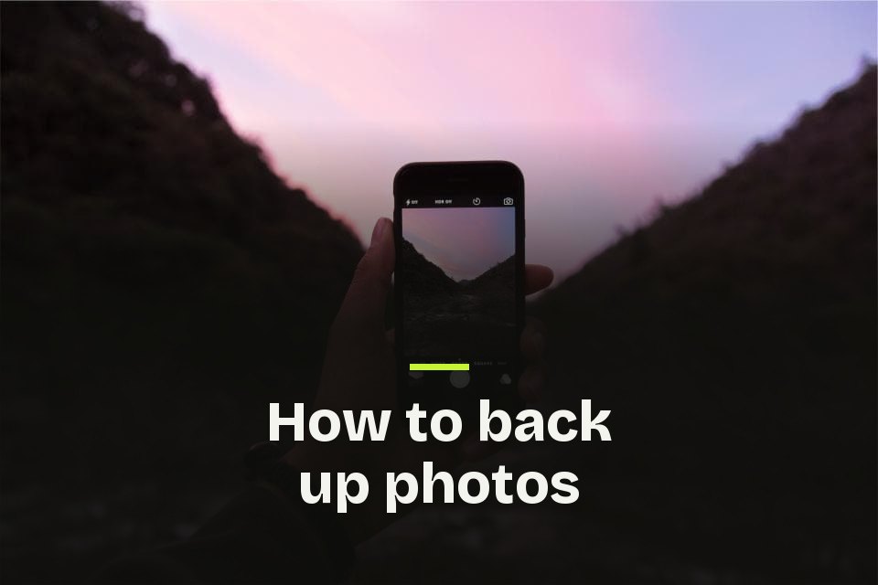 How to back up photos The words "How to back up photos" over a photo of a phone held up to photograph a mountain valley under a pink evening sky.