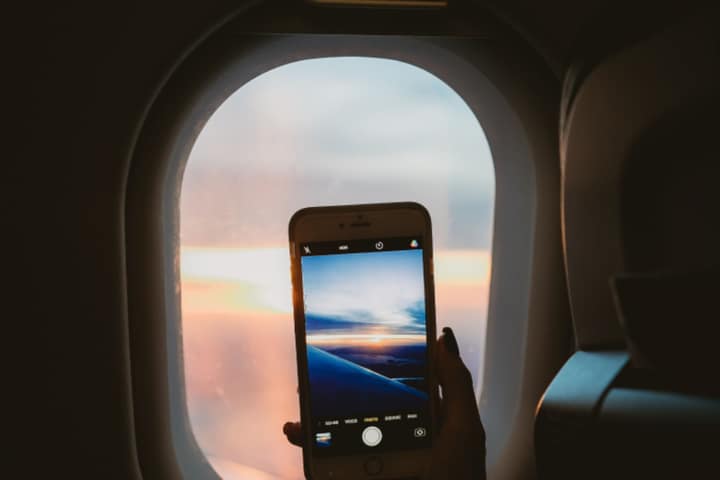 A hand holding a phone up to an airplane window at sunset, the phone's camera showing the clouds.