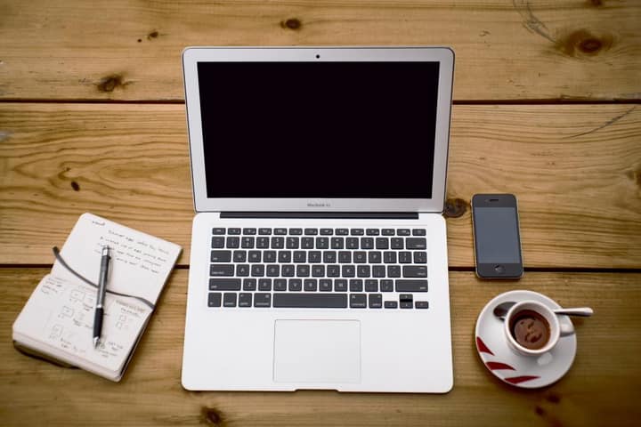 A laptop, a phone, a notebook and a coffee cup on a wooden table, seen from above.