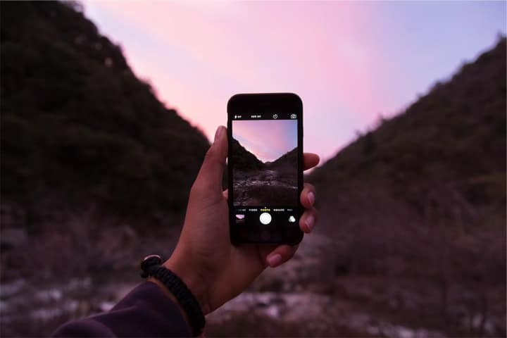 A phone held up to photograph a mountain valley under a pink evening sky.